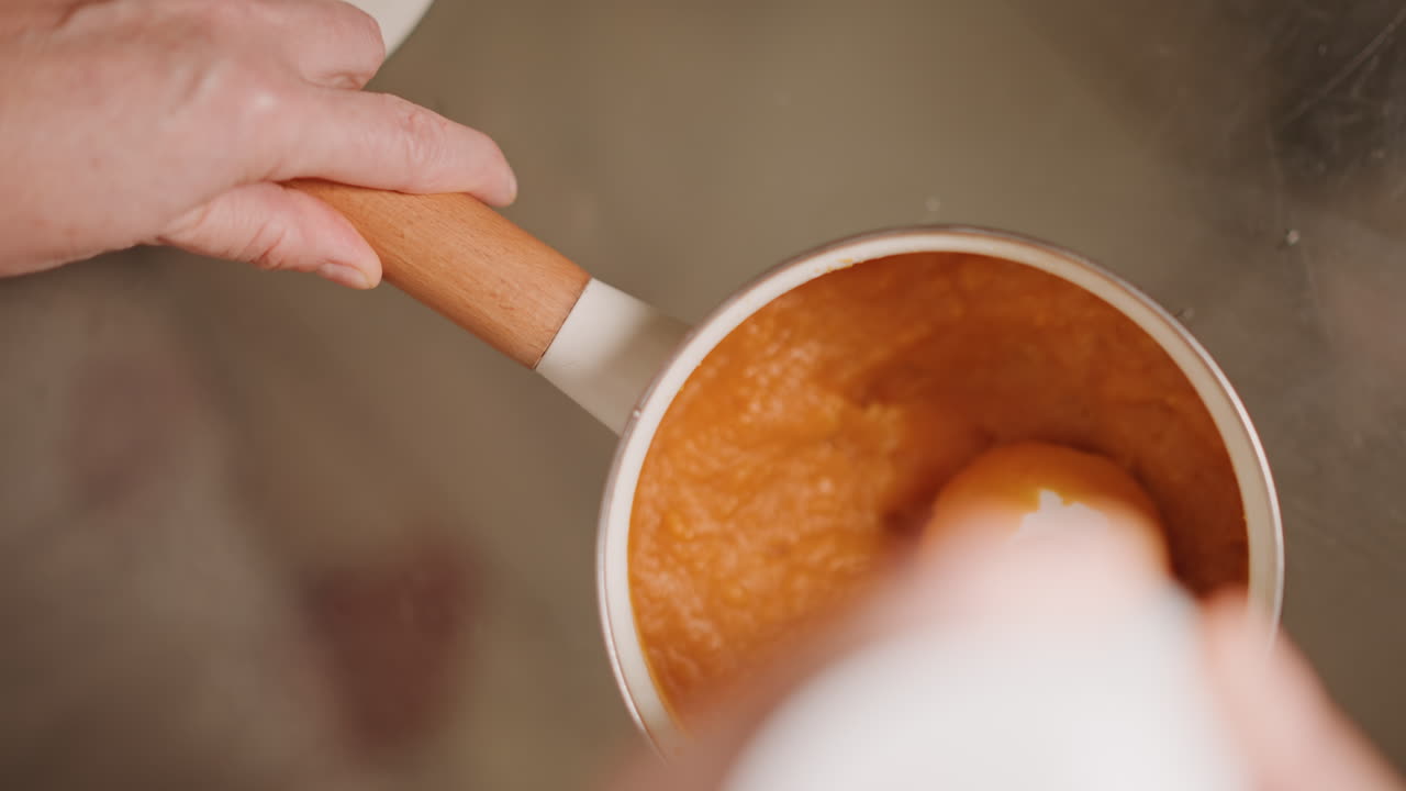 Hand blending pumpkin mixture gently in saucepan. Warm kitchen ambiance as hands blend pumpkin filling smoothly. Tactile stirring motion with immersion tool creates warm homemade pumpkin pie aroma