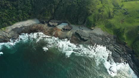 Aerial-pan-up,-black-rocky-beach,-ocean-waves-crashing-into-sea-foam