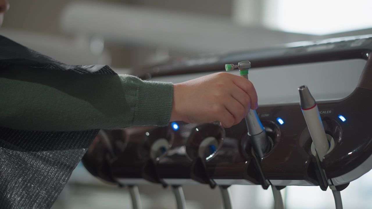 Close-up tangan pria memilih dan memegang instrumen gigi dari baki yang tertata rapi dalam pencahayaan lembut. Lengan terulur ke berbagai alat termasuk scaler dengan lampu indikator biru yang menunjukkan