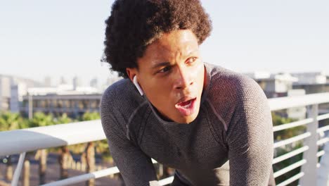 Hombre-Afroamericano-En-Forma-Haciendo-Ejercicio-Al-Aire-Libre-En-La-Ciudad,-Con-Auriculares-Inalámbricos,-Descansando-En-Una-Pasarela