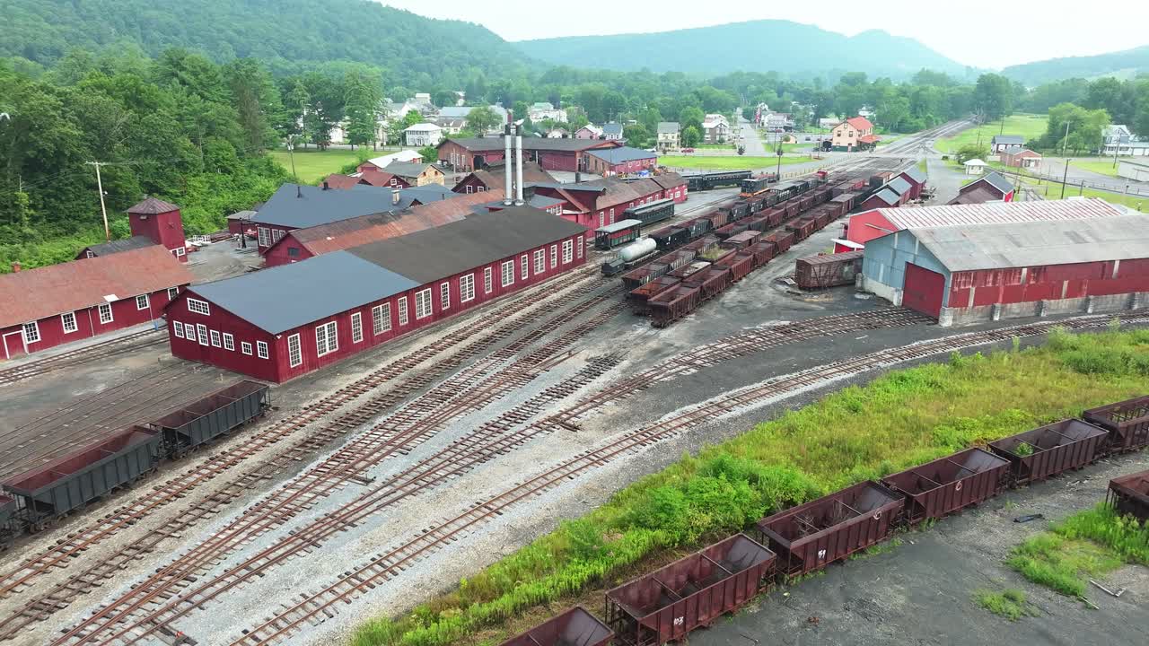 This clip showcases an aerial perspective of a train yard in a small town. Old train tracks run through the area, with storage containers, wood piles, and buildings visible in the background
