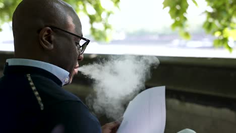 a bald, professional man vaping and reading papers while sitting on a bench beside wall