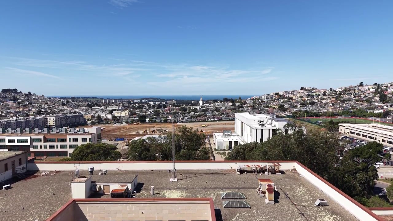Ripresa aerea con drone che inizia in un cortile soleggiato del City College di San Francisco, salendo lentamente per rivelare l'architettura dell'edificio, il campus e il paesaggio urbano circostante.