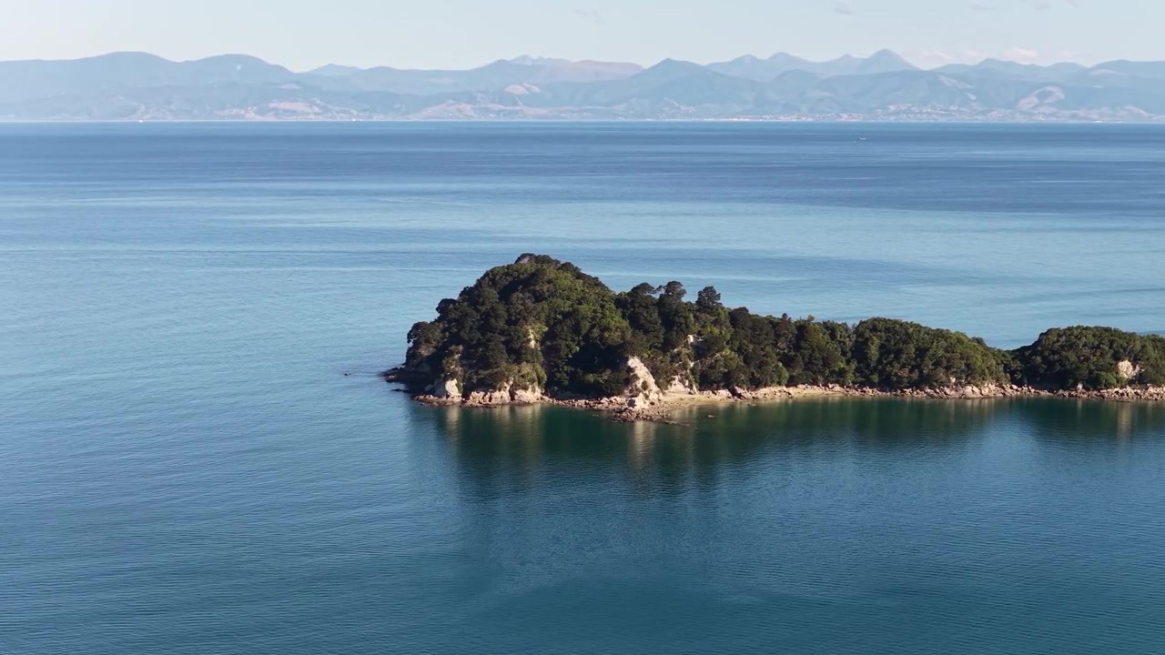 Paisaje aéreo de una isla boscosa flotando en aguas azules tranquilas frente a la Isla Sur de Nueva Zelanda. Panorama costero escénico con una atmósfera pacífica y un telón de fondo montañoso distante