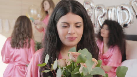 brunette woman holding and smelling a bouquet, wearing pink silk nightdress, smiling and looking at camera 1