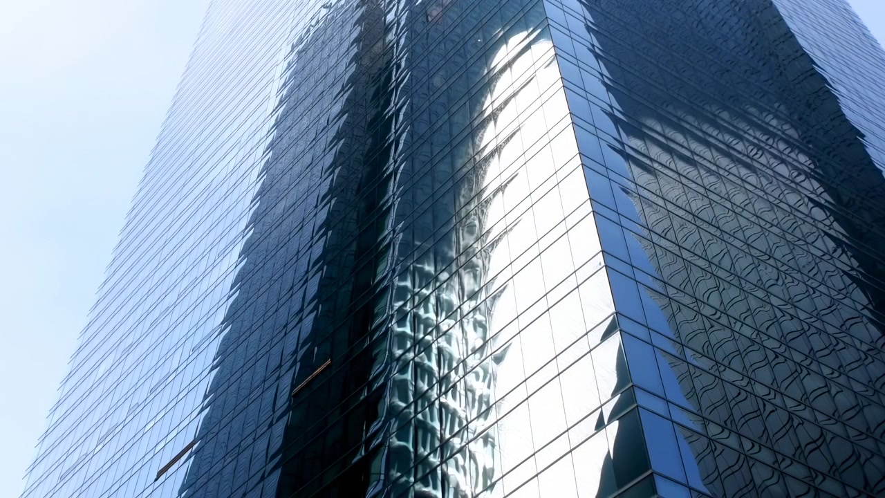 Arranha-céu alto de vidro se eleva contra o céu azul pálido, com reflexos mutáveis na fachada central escura e um brilho intenso no lado direito, criando um visual urbano corporativo limpo com forte geometria
