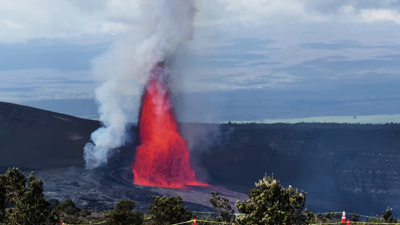 Udsigt til Kīlauea-udbruddet på Big Island, Hawaii, med en lavastråle der stiger op bag forgrundsvegetation og beskyttende pæle placeret omkring unge træer