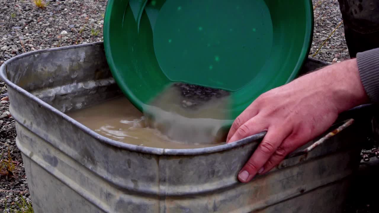 Gold panning small black fine grunge, silt, sand, and clay alluvial deposits in green trap pan, wetting, soaking, agitating and washing in tin bucket of brown murky water, static close up portrait