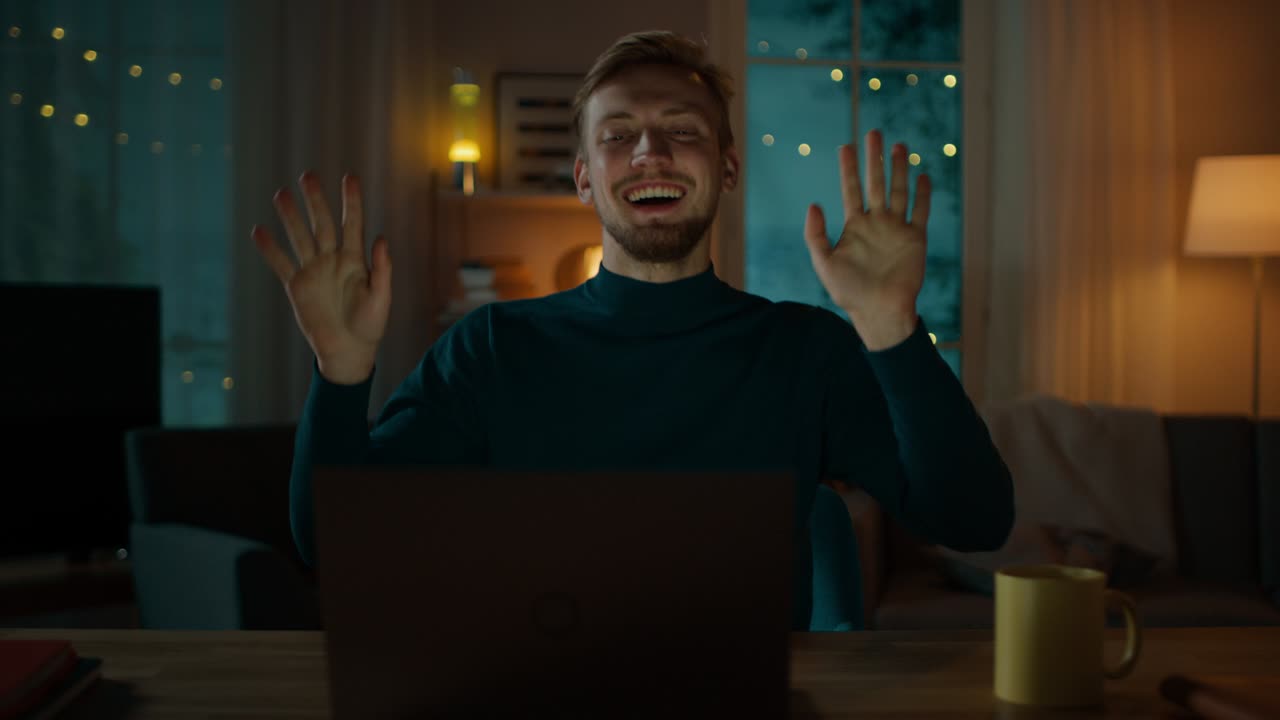 In the Evening: Happy Young Man Dances while Sitting at His Desk, He Uses Laptop and Listens to a Music.