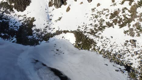 Aerial-drone-footage-above-a-beautiful-frozen-Loch-Etchachan,-before-revealing-a-dramatic-and-imposing-ice-covered-rock-face-and-cliff-with-steep-snow-filled-gullies