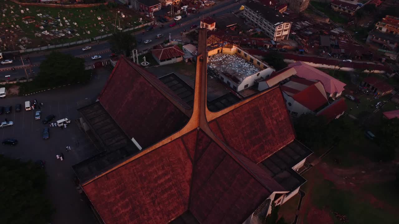 Drone video captures a unique church architectural structure from above, surrounded by an urban landscape in warm sunset light, casting long shadows and highlighting its dramatic roof design