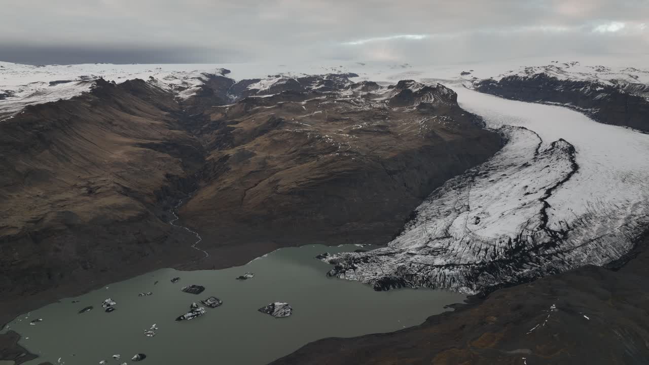 아이슬란드의 얼음산으로 가득 찬 호수로 흘러 들어가는 솔헤이마요쿨 빙하의 드론 뷰, 거친 산과 얼음으로 둘러싸인 지형
