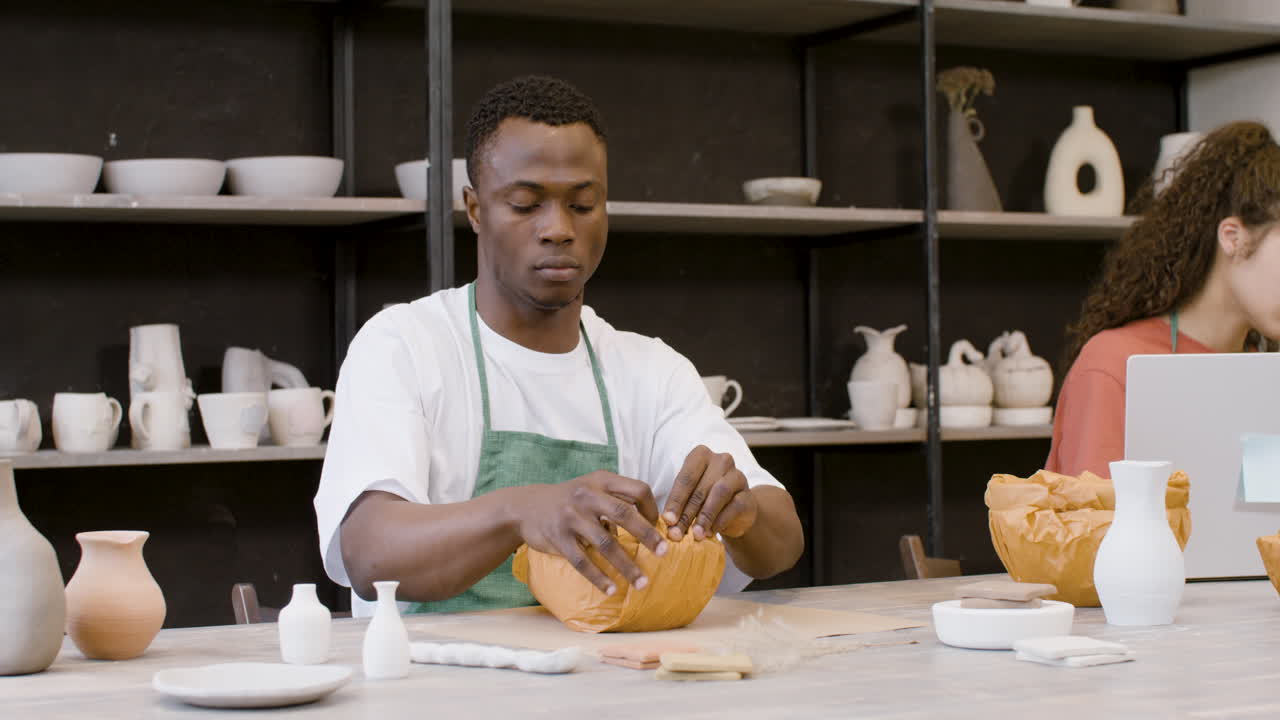 Young Man Packaging Handicraft Ceramics With Paper In The Pottery Shop While His Female Colleague Working On Laptop Computer 1
