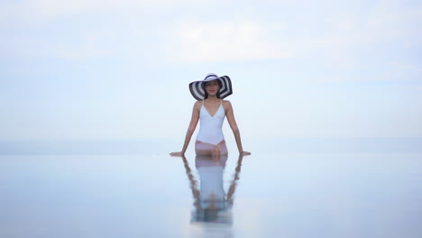 A-pretty-woman-in-a-white-one-piece-bathing-suit-and-huge,-floppy-straw-hat-giving-the-illusion-she-is-floating-in-mid-air-while-sitting-on-the-invisible-edge-of-a-resort-pool