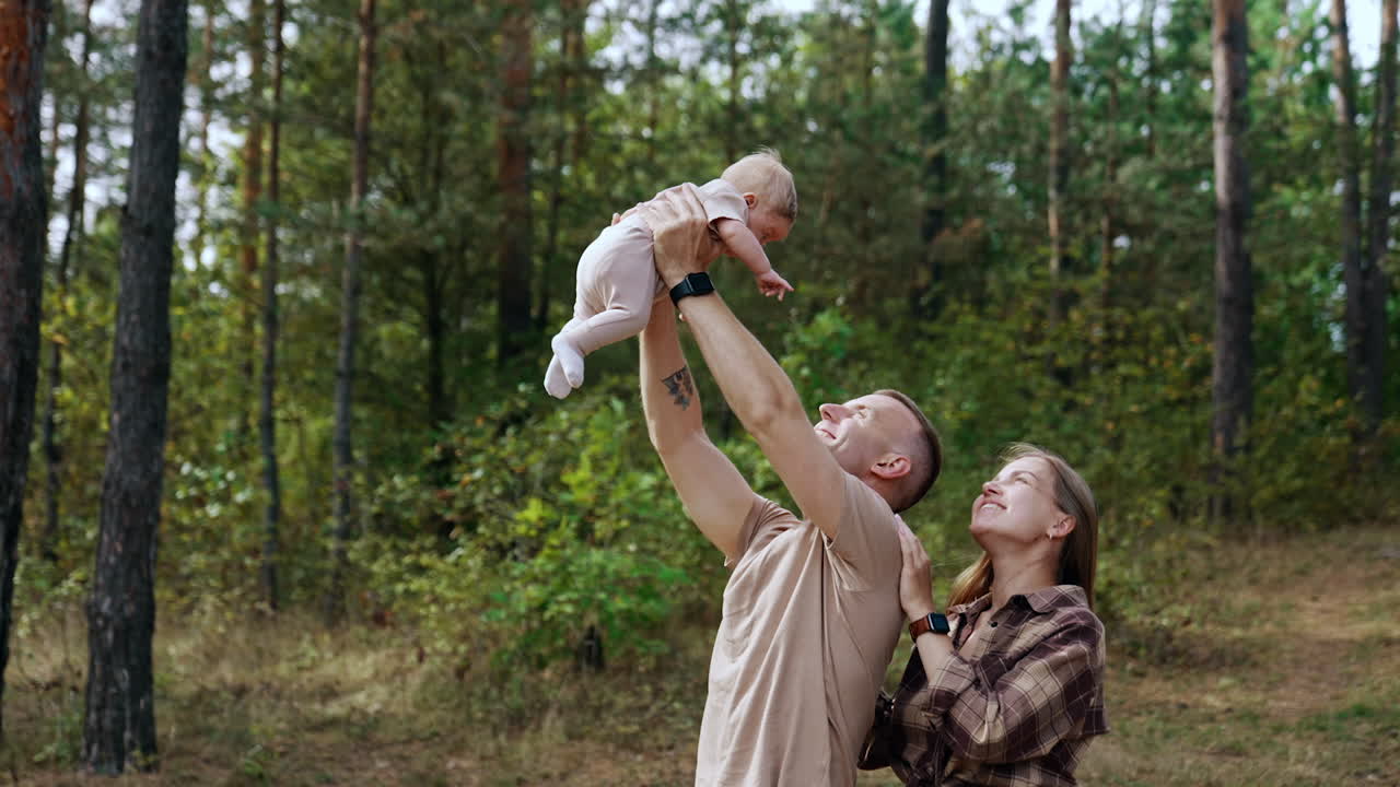 Caucasian dad raises his newborn baby above his head. Long-haired mom stands behind the man looking at child. Nature backdrop.