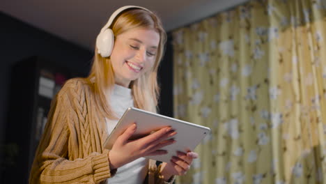 mujer joven feliz con auriculares viendo algo en la tableta mientras está de pie en la sala de estar en casa