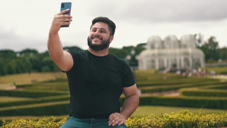 Young-man-picking-up-his-cell-phone-and-taking-a-selfie-at-the-park-outdoors-while-seated-on-a-long-wood-bench-located-at-Botanical-Garden,-Curitiba,-Brazil