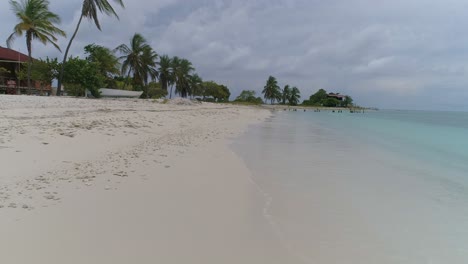 Pov-Caminando-Por-La-Impresionante-Playa-Paradisíaca-De-La-Costa,-El-Agua-Choca-Contra-La-Arena-Blanca-Y-Las-Palmeras