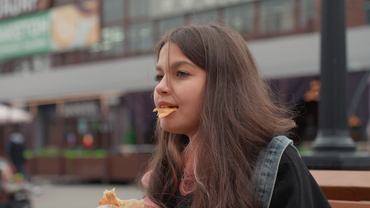 Teen girl with scarf and backpack. Young woman seated on bench with pastry and pensive look. Girl in autumn attire delicately eating pastry with thoughtful expression at urban market setting
