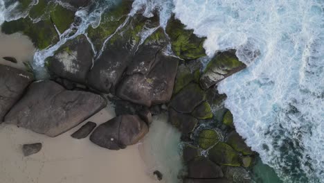 aerial top down view: waves crash against rocks along coastline and sandy beach during cloudy day