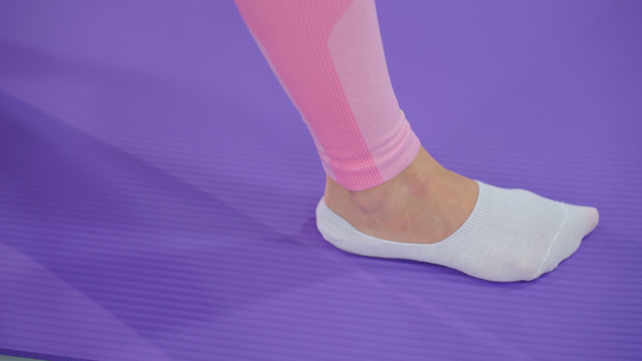색 양말과 분홍색 레을 입고 요가 매트에 어있는 다리의 클로즈업, 운동에 참여하고, 부드러운 보라색 배경으로 움직이는 손의 부분적 인 시각