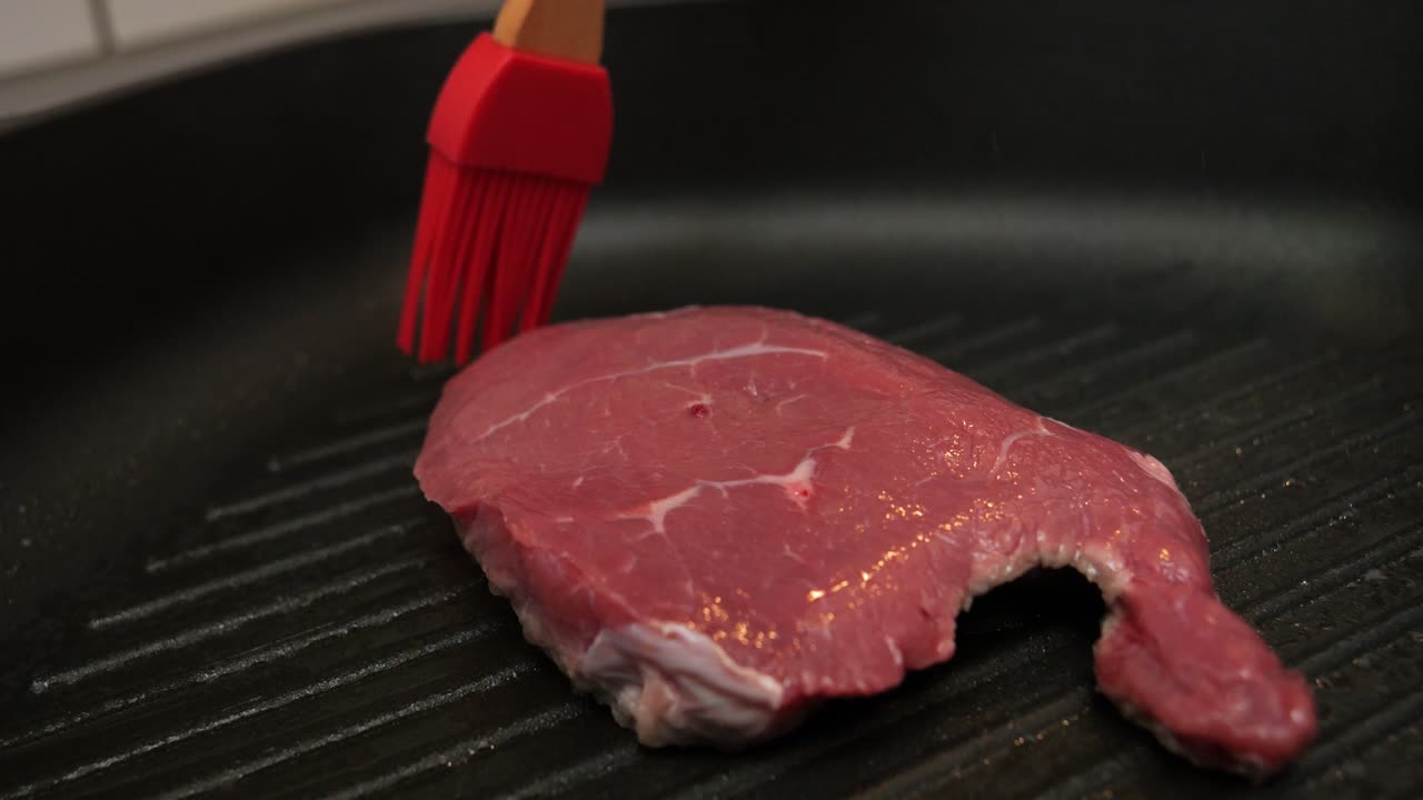 맛있는 저녁 식사를 위해 부엌에 있는 뜨거운 팬에 있는 쇠고기 스테이크를 기름으로 닦는 빨간 붓의 클로즈업 샷