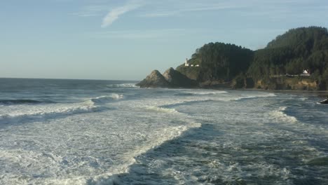 wide aerial moving low across waves toward haceta head lighthouse in oregon