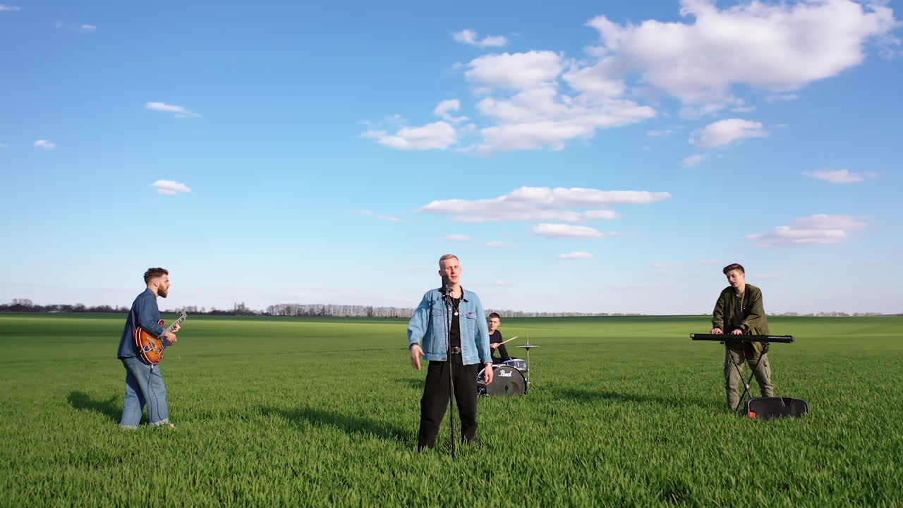 Blond vocalist sings song at microphone stand in the meadows. Three men play musical instruments at backdrop. Circle movement shot.