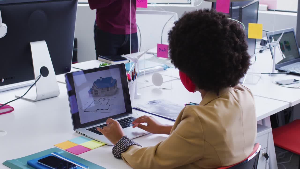 mujer de negocios de raza mixta con máscara facial sentada usando una computadora portátil en una oficina moderna