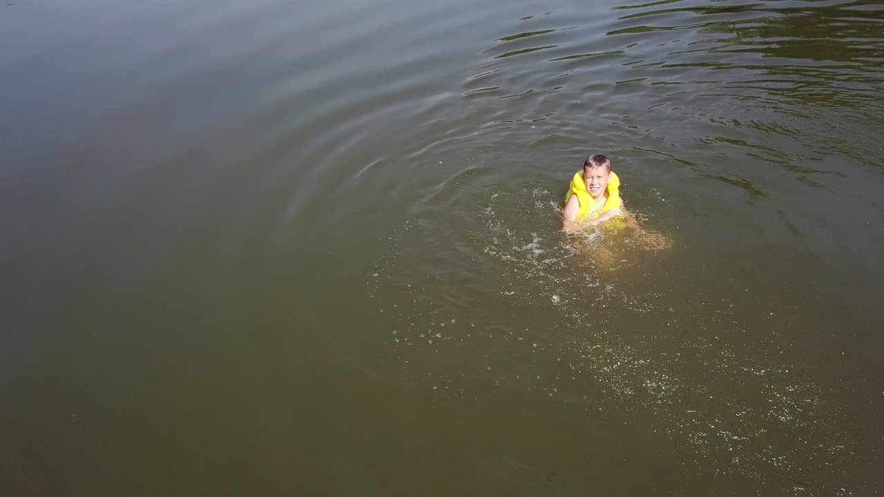 Little kid cheerfully swimming in the countryside isolated in the water background. Boy wearing vest to be safe swims happily and makes splashes in water. Aerial view