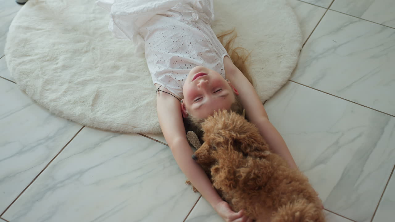 Girl in white gown lying on white rug, playing with her dog, enjoying playful moments together at home, bonding with furry companion in cozy environment