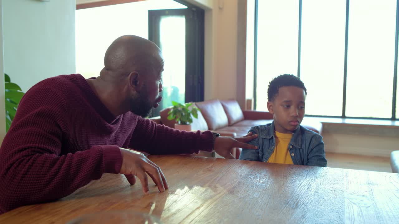 African American father and son at table speaking as son covering ears, father comforting shoulder