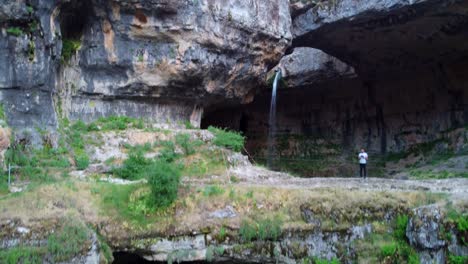 waterfall behind a natural bridge covered in lush green vegetation, baatara gorge waterfall in lebanon - drone ascending