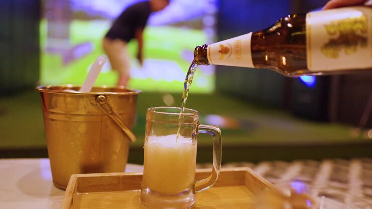 Una persona versa birra in un boccale presso un simulatore di golf al coperto con illuminazione soffusa.