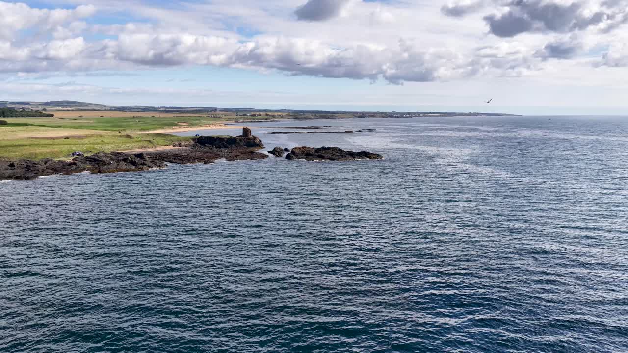 Drone glides toward rugged Fife coastline, ancient stone ruins, and calm ocean under cloudy daylight