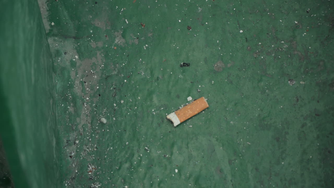 Cigarette butt on green pavement aerial drone view shows discarded filter among small debris and stains near sidewalk edge closeup texture highlights ash nicotine residue and environmental hazard