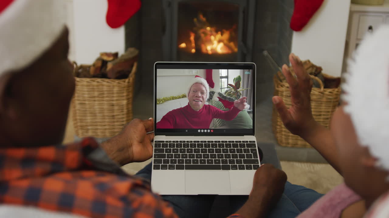 Coppia afroamericana con cappelli di Babbo Natale che utilizza un laptop per una videochiamata di Natale con un uomo sullo schermo.