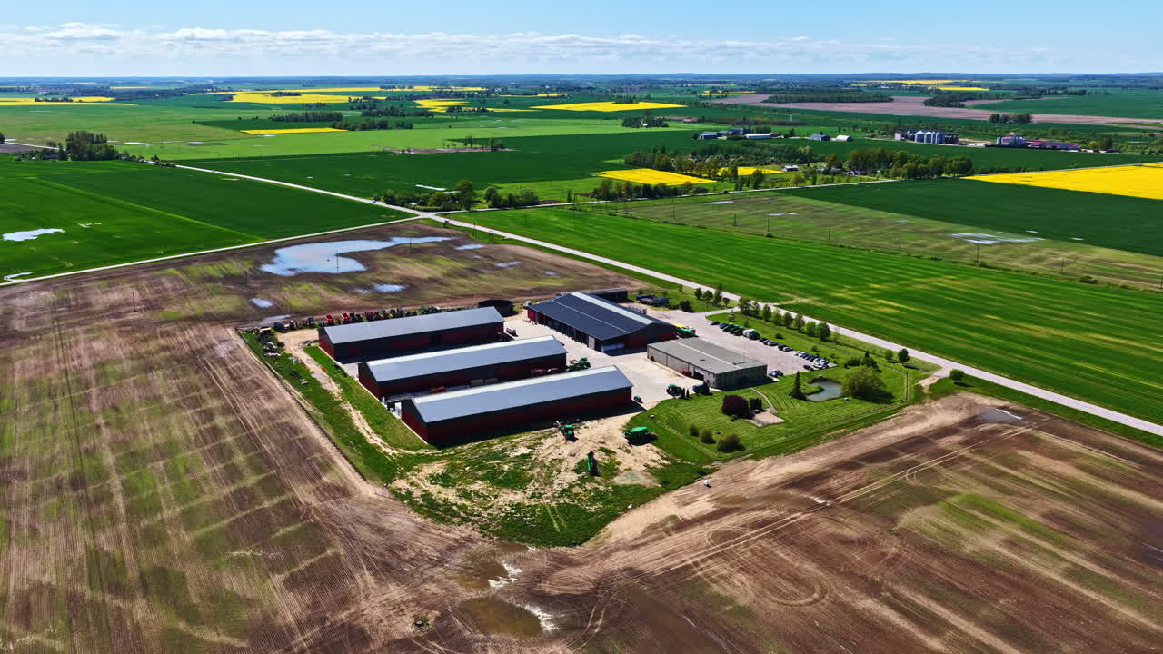 Aerial view of a large farm facility surrounded by wide agricultural fields, with storage barns, access roads, green crop areas, and patches of soil after recent rain, captured in bright daytime