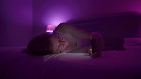 girl laying on side browses social media on smart phone late at night in bedroom.