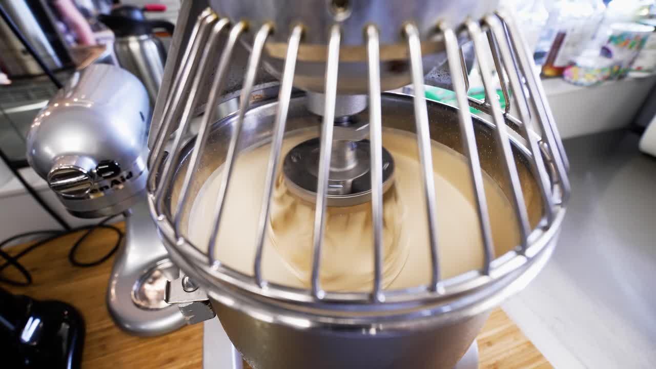 Stainless steel commercial stand mixer rapidly whisks batter, numerous stand mixers of various sizes clutter bakery kitchen counter, slow motion 4K