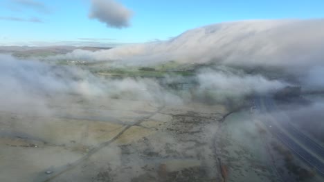 Flying-towards-large-hill-covered-by-drifting-thick-cloud-and-foreground-drifting-mist-with-M6-motorway-beside-at-dawn-in-winter