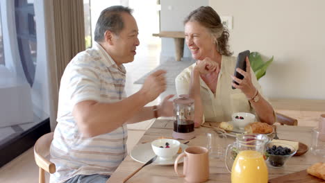 smiling senior couple enjoying breakfast and looking at smartphone together