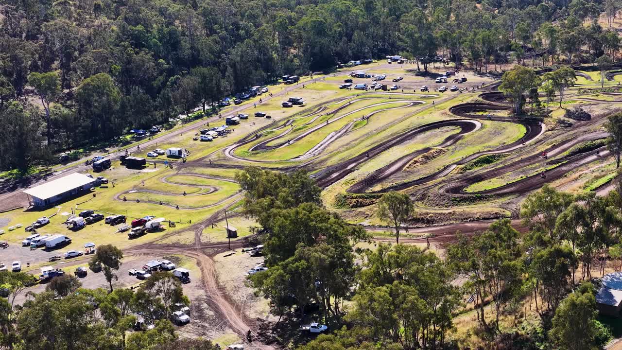 Drone pans over motocross track, parked vehicles, and forested landscape in bright daylight