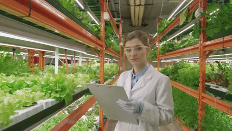 Junge-Gewächshausarbeiterin-In-Weißem-Kittel-Und-Brille,-Die-Klemmbrett-Hält,-Während-Sie-In-Die-Kamera-Innerhalb-Der-Vertikalen-Farm-Schaut