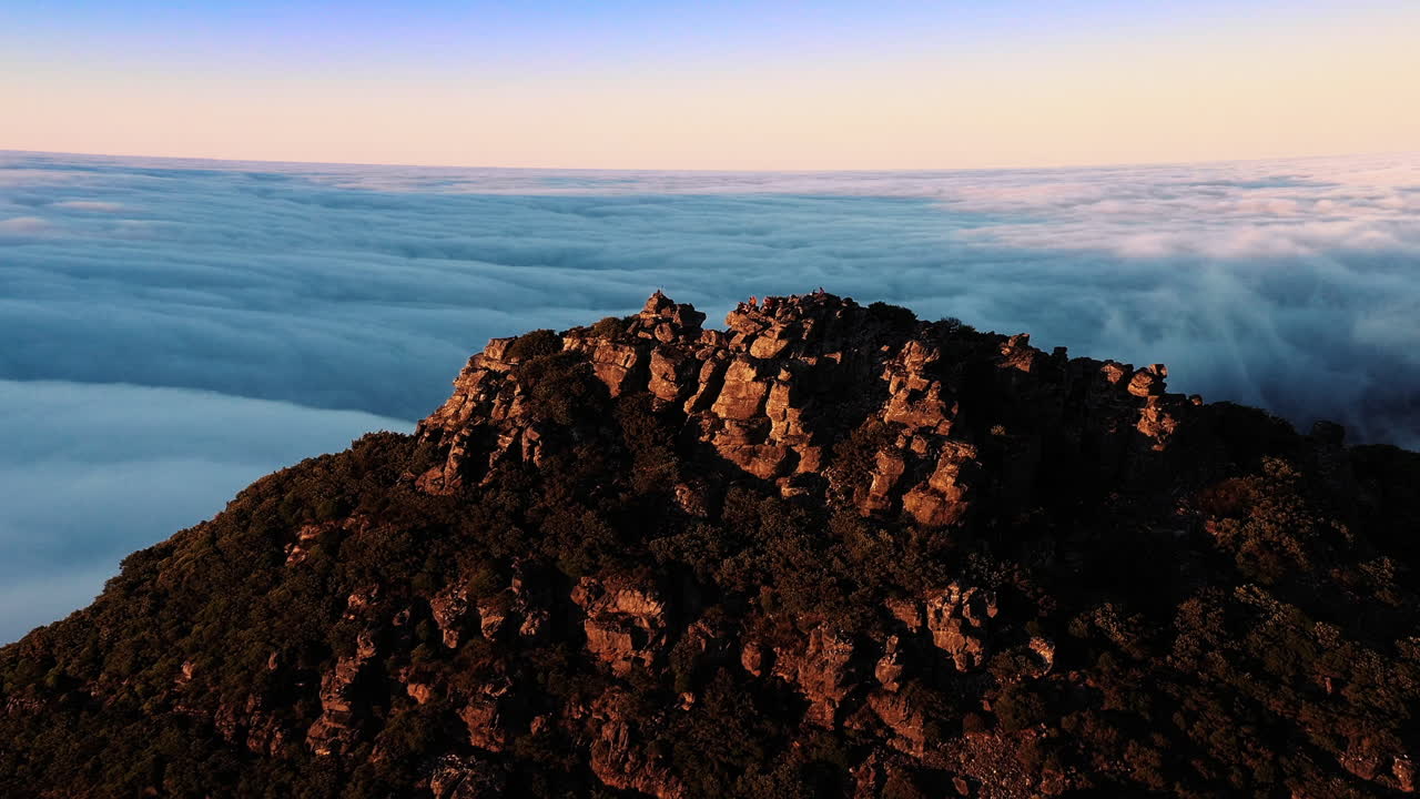 Aerial, drone view around a group of hikers, standing on a sunlit summit of a rocky mountain, surrounded by clouds, during golden hour, in Hassell National Park, in West Australia