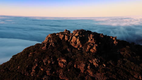 aerial, drone view around a group of hikers, standing on a sunlit summit of a rocky mountain, surrounded by clouds, during golden hour, in hassell national park, in west australia
