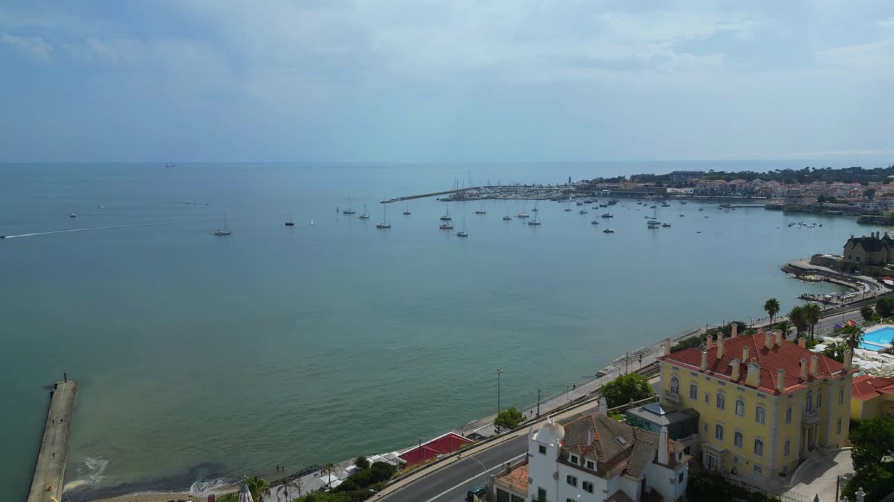 Volando sopra le case verso il mare con la baia di Cascais e il suo porto turistico con barche ormeggiate sullo sfondo.