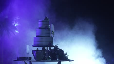 Dramatically-lit-wedding-cake-with-floral-decorations,-surrounded-by-mist-and-purple-lighting