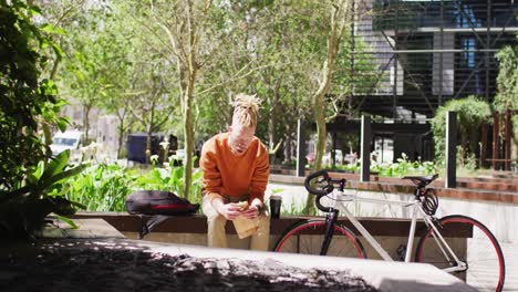 albino african american man with dreadlocks sitting in park with bike eating sandwich