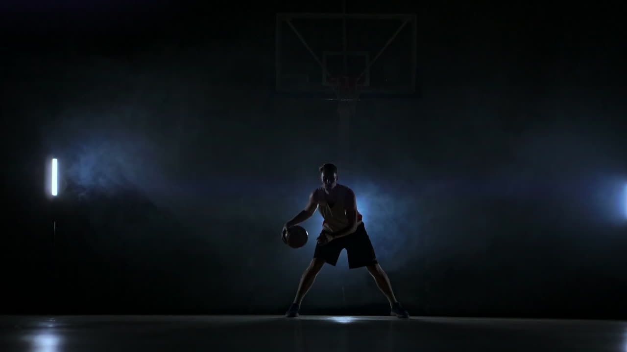 Skill dribbling basketball player in the dark on the basketball court with backlit back in the smoke. Slow motion streetball